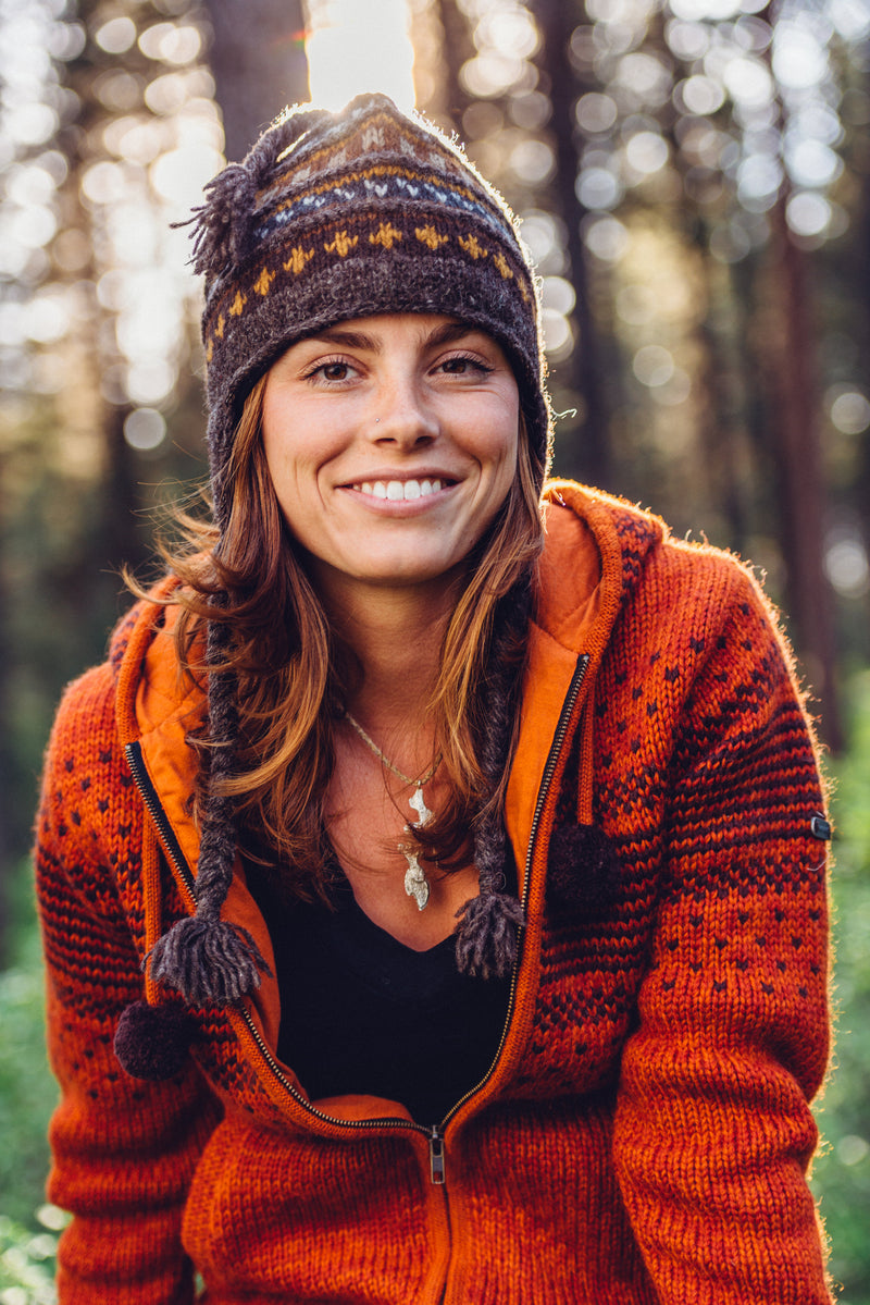 Woman wearing Everest Designs brown Sherpa Earflap in Ale and the zip-up Ghandaki sweater in Orange while seated in the woods